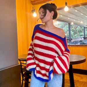 May include: A red, white, and blue striped sweater with a wide neckline. The sweater has long sleeves and a loose fit. The neckline and cuffs are trimmed with blue. The sweater is paired with blue jeans.