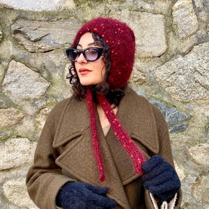 Könnte beinhalten: Eine bordeauxfarbene Strickmütze mit roten Pailletten und langen Bändern. Die Mütze wird mit einer schwarzen Sonnenbrille, einem braunen Mantel und dunkelblauen Handschuhen getragen. Es ist ein Winteraccessoire.
