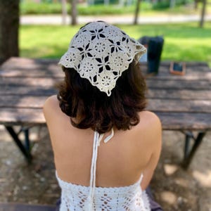 May include: A cream-colored, crocheted headscarf with a floral pattern. The head covering is square-shaped and tied at the back. The person is wearing a matching crocheted halter top. The background is a wooden table and a park setting.