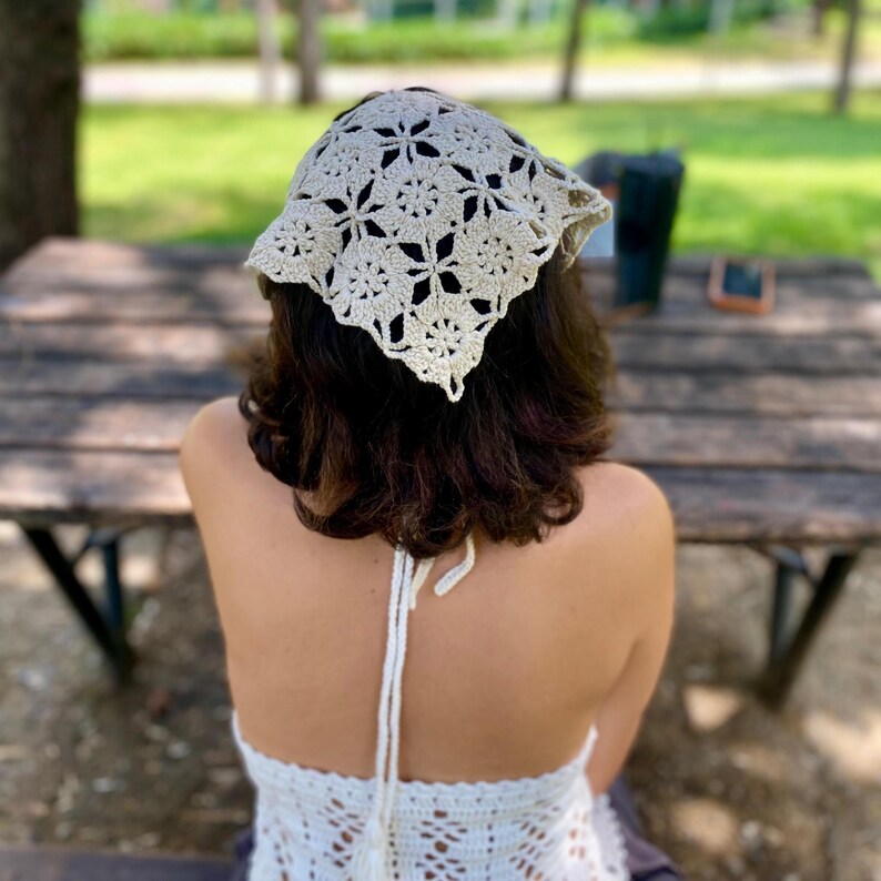 May include: A cream-colored, crocheted headscarf with a floral pattern. The head covering is square-shaped and tied at the back. The person is wearing a matching crocheted halter top. The background is a wooden table and a park setting.