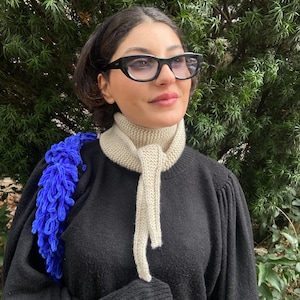 May include: A black sweater with a cream-colored knitted scarf and a blue shoulder bag. The scarf is tied in a knot. The person is wearing black-framed glasses.