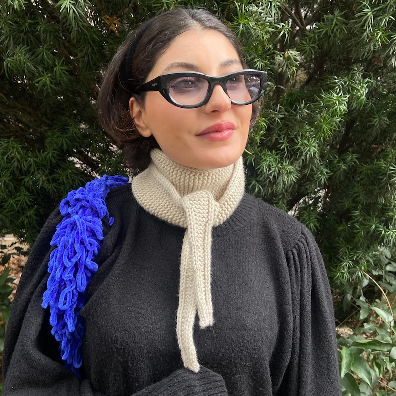 May include: A black sweater with a cream-colored knitted scarf and a blue shoulder bag. The scarf is tied in a knot. The person is wearing black-framed glasses.