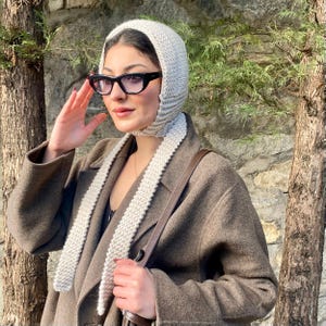 May include: A light gray knitted hood with long scarf-like ties. The hood is worn over the head and drapes down the front. The person is wearing black framed glasses and a brown coat. The background is a stone wall and tree.