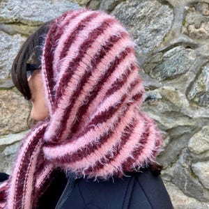 Puede incluir: Conjunto de capucha y bufanda a rayas, en tonos rosa y burdeos. El accesorio de punto tiene un aspecto suave y texturizado con rayas alternas. La bufanda est&aacute; sobre una chaqueta negra, creando un look acogedor y elegante.