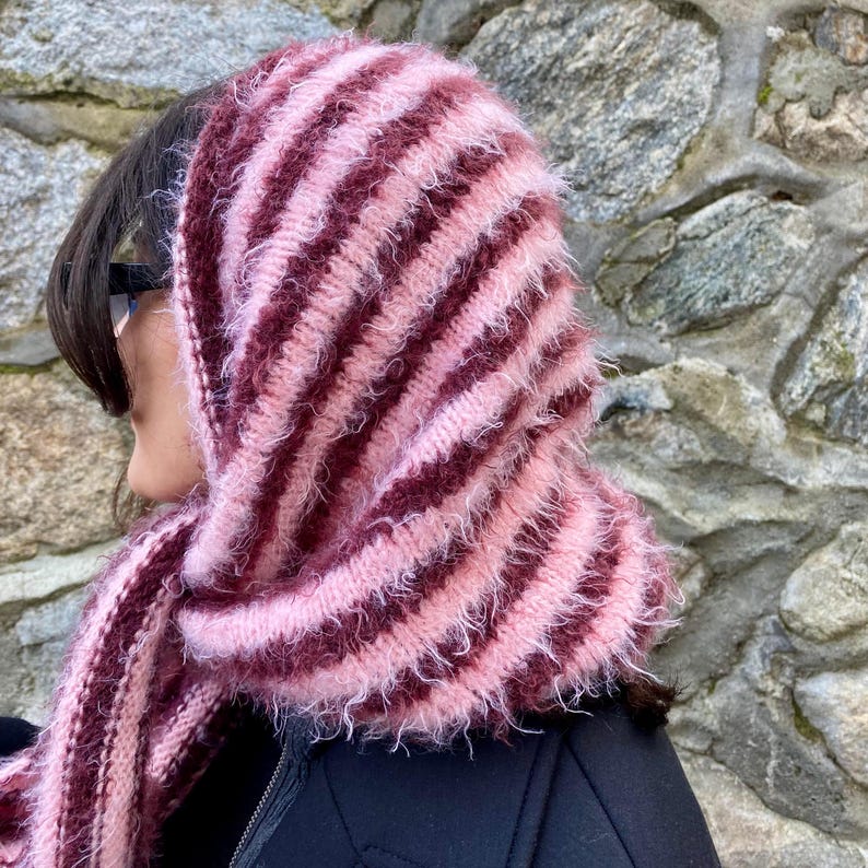 Puede incluir: Conjunto de capucha y bufanda a rayas, en tonos rosa y burdeos. El accesorio de punto tiene un aspecto suave y texturizado con rayas alternas. La bufanda est&aacute; sobre una chaqueta negra, creando un look acogedor y elegante.