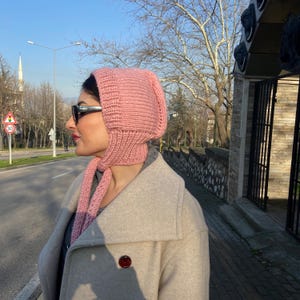 May include: A pink knitted hat with a long scarf attached. The hat has a pointed top and covers the ears. The scarf is folded in half and hangs down the front of the person wearing it.