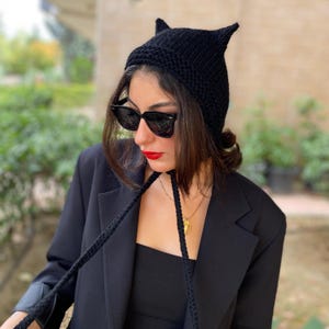 May include: A black knit hat with pointed ears. The hat has long ties and is worn with sunglasses and a black blazer. The hat is a unique accessory, perfect for adding a touch of whimsy to any outfit.