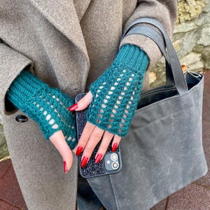 May include: Teal fingerless gloves with a knitted pattern. The gloves are worn with a grey coat and a grey tote bag. The person is holding a black phone with a glittery case. The nails are painted red.