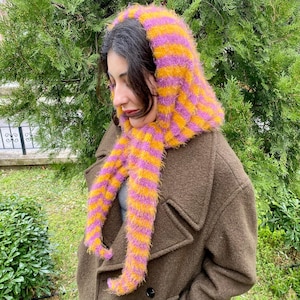 Puede incluir: Conjunto de capucha y bufanda a rayas y esponjoso en tonos amarillo, naranja y morado. La bufanda cuelga por delante de un abrigo marr&oacute;n. La capucha cubre la cabeza y el cuello, con la bufanda extendi&eacute;ndose por el pecho.