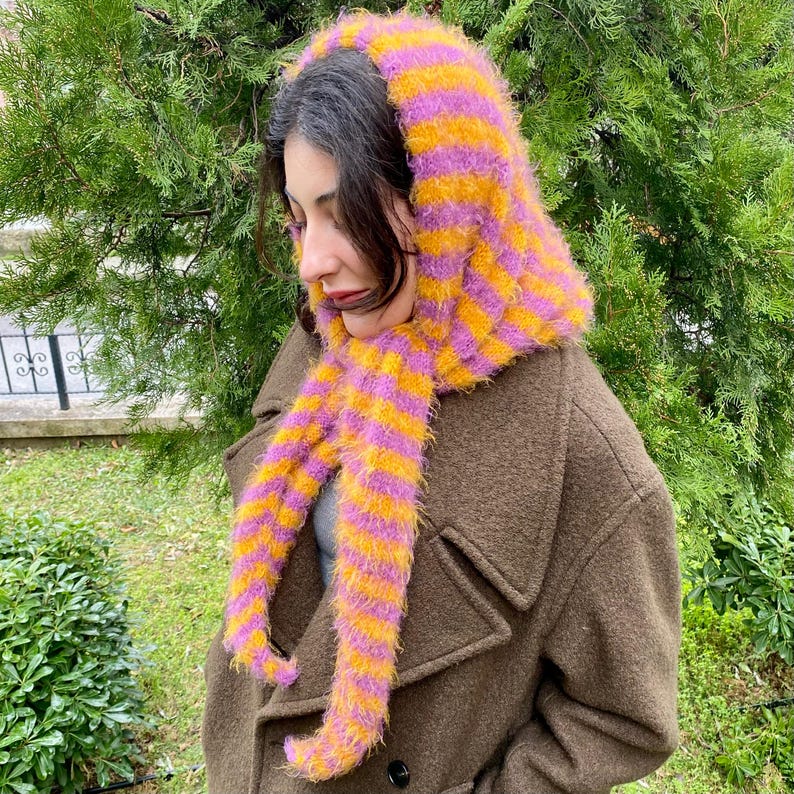 Puede incluir: Conjunto de capucha y bufanda a rayas y esponjoso en tonos amarillo, naranja y morado. La bufanda cuelga por delante de un abrigo marr&oacute;n. La capucha cubre la cabeza y el cuello, con la bufanda extendi&eacute;ndose por el pecho.