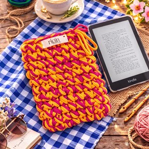 May include: A handmade, yellow and pink crocheted Kindle sleeve with the brand name "rioin" on a tag. The sleeve rests on a blue and white checkered cloth, next to a Kindle e-reader, a cup of coffee, and other craft supplies.