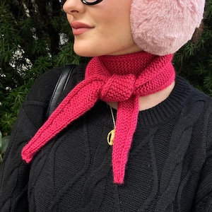 May include: A bright pink knitted scarf tied around the neck. The scarf is made of a thick yarn and has a textured appearance. The person is wearing a black sweater and pink earmuffs. A gold necklace is visible.