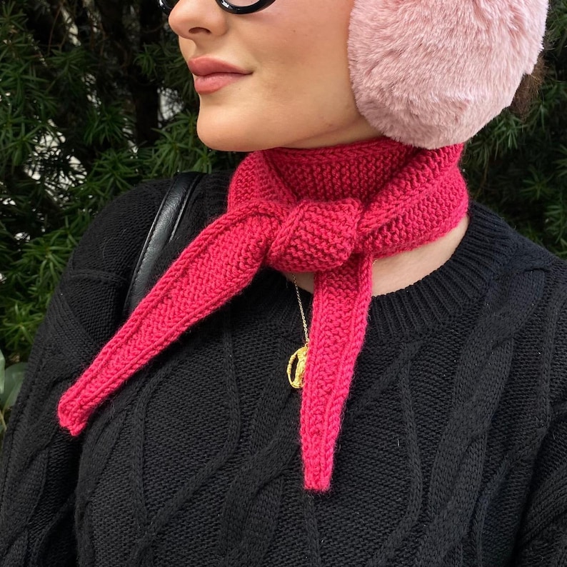 May include: A bright pink knitted scarf tied around the neck. The scarf is made of a thick yarn and has a textured appearance. The person is wearing a black sweater and pink earmuffs. A gold necklace is visible.