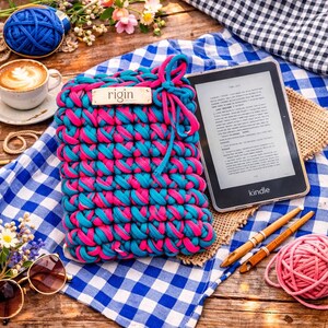 May include: A handmade, crochet Kindle sleeve in vibrant pink and teal. The sleeve features the word "rigin" on a wooden tag. A Kindle e-reader rests beside the sleeve on a blue and white checkered cloth. Other items include yarn, a cup of coffee, and sunglasses.