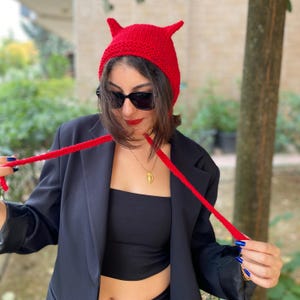 May include: A bright red knit hat with two pointed horns. The hat has long red ties. The person in the image is wearing a black blazer, black top, and sunglasses. The hat is a unique accessory.