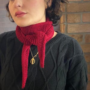 May include: A knitted red scarf with a pointed end, worn around the neck. A gold-colored pendant hangs from a delicate chain. The scarf is made of a thick yarn, and the overall style is cozy and handmade.