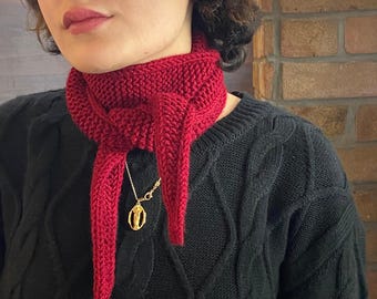 May include: A knitted red scarf with a pointed end, worn around the neck. A gold-colored pendant hangs from a delicate chain. The scarf is paired with a black cable-knit sweater.