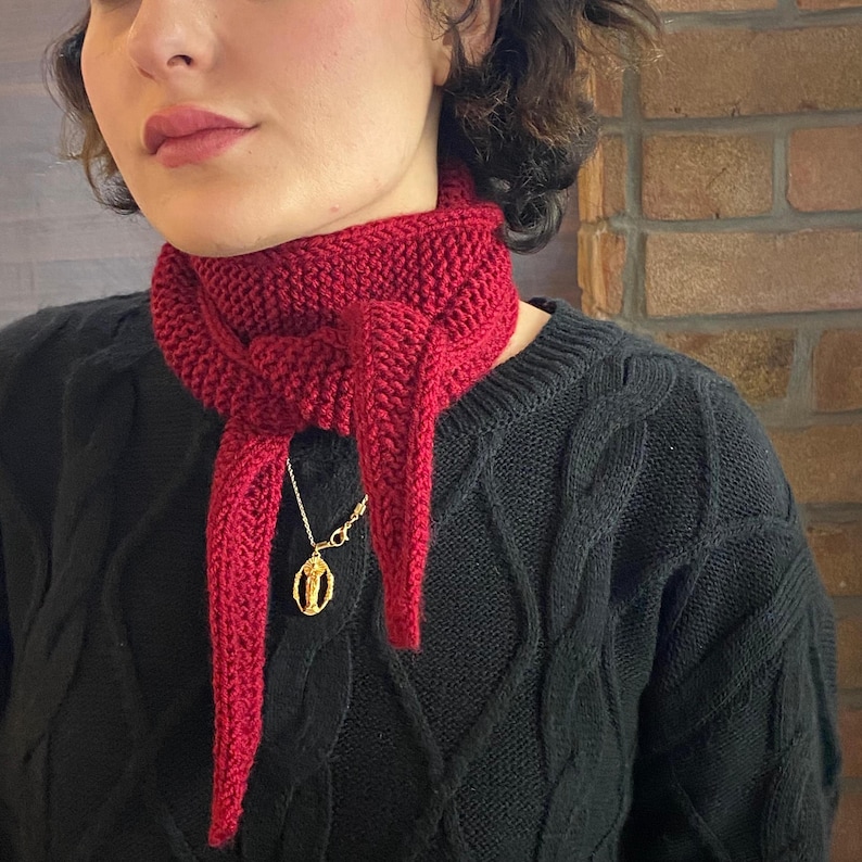 May include: A knitted red scarf with a pointed end, worn around the neck. A gold-colored pendant hangs from a delicate chain. The scarf is made of a thick yarn, and the overall style is cozy and handmade.