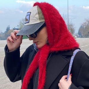May include: A bright red fuzzy knit headscarf worn over a beige and grey baseball cap with a patch on the front. The cap features a dark grey brim and a light beige crown. The headscarf is textured with long fibers and ties under the chin.