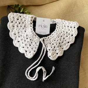 Może przedstawiać: Biały koronkowy kołnierzyk z zawiązywaniem. Kołnierzyk leży na czarnym swetrze. Etykieta na kołnierzyku głosi "Studio rigin".