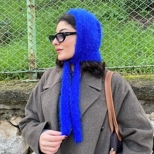 Może przedstawiać: Żywa niebieska dzianinowa kominiarka z długimi wiązaniami. Czapka wykonana jest z puszystego materiału i jest noszona z szarym płaszczem. Tło przedstawia zielone ogrodzenie i liście. Osoba nosi czarne okulary przeciwsłoneczne.