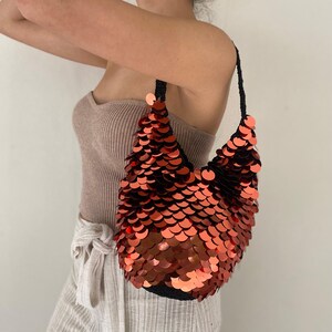 May include: A hobo-style shoulder bag covered in overlapping, metallic copper-colored circular sequins. The bag features a black crocheted base and a matching black crocheted shoulder strap, creating a distinct contrast with the shiny, reflective surface.