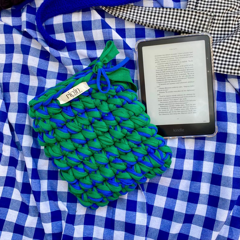 May include: A green and blue knitted pouch with a drawstring closure, labeled "rigin", rests on a blue and white checkered fabric. A Kindle e-reader with a clear case is also present, displaying text on its screen.