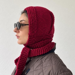 Könnte beinhalten: Eine dunkelrote Stricksturmhaube mit dickem, strukturiertem Rand und langen, integrierten Schalbindern. Die Kopfbedeckung weist ein grobes Strickmuster auf und wird über einer braunen Steppjacke getragen, kombiniert mit einer dunklen Sonnenbrille.