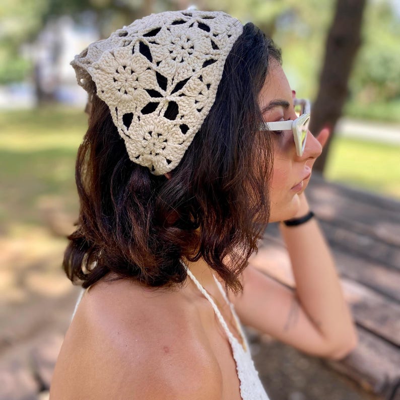 May include: A beige crochet bandana with a floral pattern. The head covering is triangular and worn on the head. The bandana is made of a soft, textured yarn. The person is wearing white sunglasses and a white top.