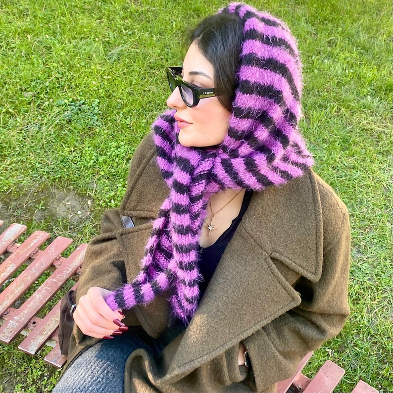 Puede incluir: Un conjunto de capucha y bufanda de punto a rayas moradas y negras. La capucha est&aacute; puesta sobre la cabeza y la bufanda est&aacute; drapeada alrededor del cuello. La persona lleva un abrigo verde oscuro y gafas de sol.