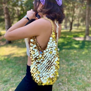 May include: A shoulder bag crafted from tan crochet yarn and densely covered in reflective, circular gold sequins. The bag features a single integrated crochet strap and a slouchy, unstructured silhouette that hangs vertically from the shoulder.