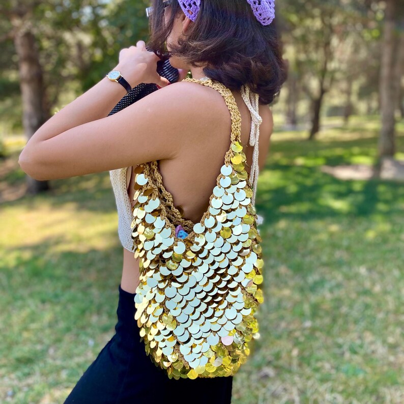 May include: A shoulder bag crafted from tan crochet yarn and densely covered in reflective, circular gold sequins. The bag features a single integrated crochet strap and a slouchy, unstructured silhouette that hangs vertically from the shoulder.