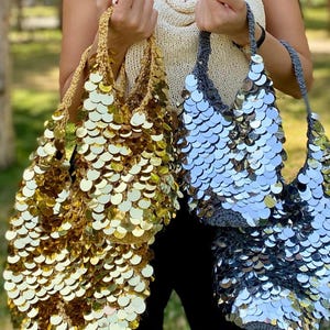 May include: Two hobo-style bags made of crocheted yarn covered in large, reflective circular sequins. One bag is gold with gold sequins, and the other is gray with silver sequins. Both bags feature a single handle and a slouchy, unstructured shape.