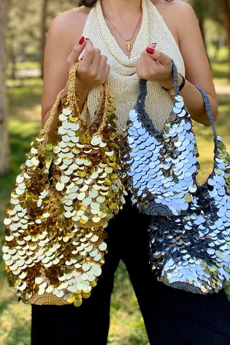 May include: Two hobo-style bags made of crocheted yarn covered in large, reflective circular sequins. One bag is gold with gold sequins, and the other is gray with silver sequins. Both bags feature a single handle and a slouchy, unstructured shape.