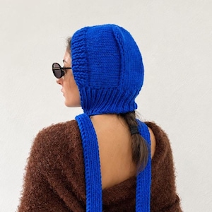 Könnte beinhalten: Eine leuchtend blaue, gestrickte Sturmhaube mit einer dicken Rippenstruktur um die Gesichtsöffnung und zwei langen, passenden blauen Strickbändern, die von der Basis der Kapuze herabhängen, gefertigt aus grobem Garnmaterial.