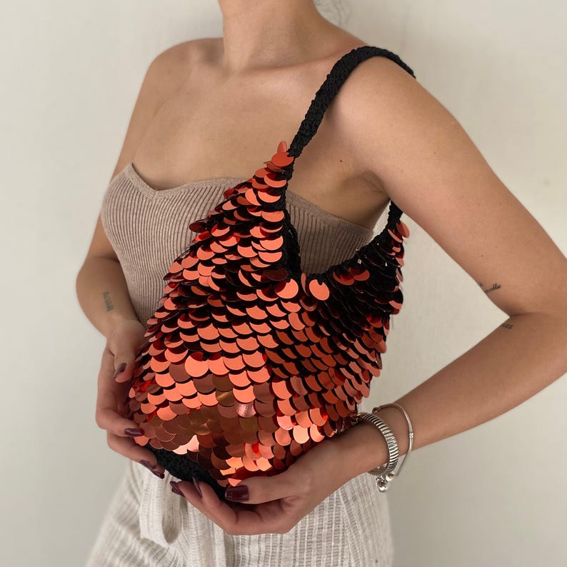 May include: A hobo-style shoulder bag covered in overlapping, reflective orange-red circular sequins. The bag features a black crocheted base and a matching black crocheted shoulder strap, creating a textured, scale-like appearance across the surface.