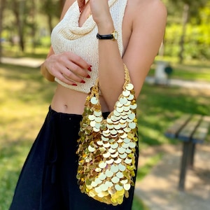 May include: A small handbag crafted from a crocheted base and covered in overlapping, reflective gold-toned circular sequins. The bag features a soft, unstructured shape with a single integrated handle, creating a shimmering, metallic appearance.