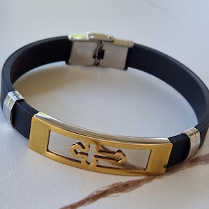 May include: Black rubber bracelet with a gold and silver cross design. The bracelet features a stainless steel clasp and accents. The cross is centered on a rectangular gold plate with a silver background.