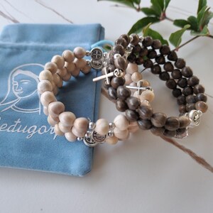 May include: Three wooden beaded bracelets with silver charms. The bracelets are stacked on top of each other and are in a blue velvet pouch. The pouch has the word "Medugorje" embroidered on it.