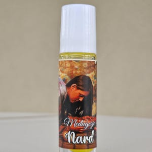May include: A clear glass roll-on bottle with a white cap. The label features an image of a person and the words "Medjugorje Nard" in elegant script. The bottle contains a yellow liquid.