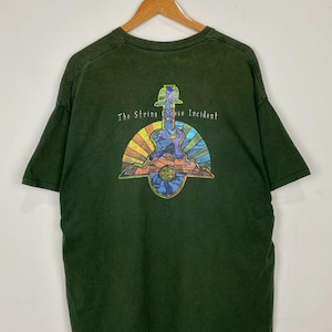 May include: Dark green t-shirt with a colorful graphic featuring a guitar, sun, and mountain design. The text "The String Cheese Incident" is arched above the graphic. The shirt is hanging on a wooden hanger.