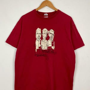May include: A red t-shirt featuring a graphic of three sketched faces. The text "VIOLENT FEMMES" is printed below the faces. The t-shirt is hanging on a wooden hanger against a white background.
