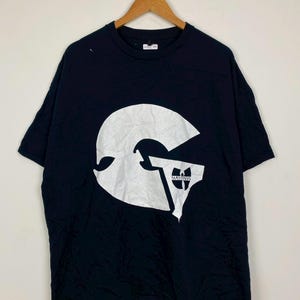 Puede incluir: Camiseta negra de manga corta con un gran gráfico blanco en la parte delantera. El gráfico es una 'W' estilizada con un logotipo más pequeño debajo. La camiseta está colgada en una percha de madera sobre un fondo liso.