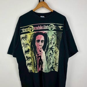 Puede incluir: Camiseta negra con un gráfico colorido de una persona con rastas fumando. El diseño incluye el texto "Excuse me while I light my spliff". El diseño también incorpora imágenes de leones y hojas de cannabis.
