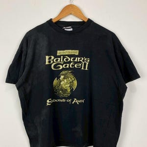 May include: Black t-shirt featuring the text "Baldur's Gate II" in gold lettering, with a gold graphic of a winged figure. The shirt is hanging on a wooden hanger.