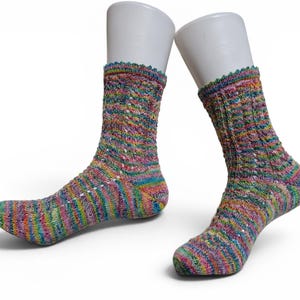 Op de afbeelding: Een paar halflange gebreide sokken met een levendig, meerkleurig gemêleerd garenpatroon. De sokken hebben een gestructureerd kabelbreisel met kleine gaatjesdetails en een decoratieve picotrand aan de boord, gepresenteerd op witte vormen.