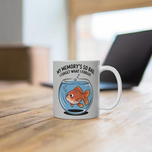 Op de afbeelding: Witte keramische mok met een cartoon goudvis in een kom en de tekst "MY MEMORY'S SO BAD, I FORGET WHAT I FORGOT." De goudvis is oranje en heeft een verdrietige uitdrukking. De mok staat op een houten oppervlak.