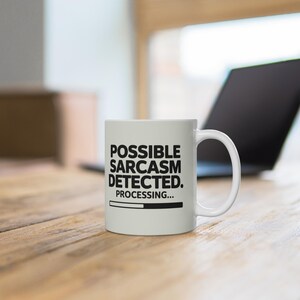 Op de afbeelding: Witte keramische mok met de tekst "POSSIBLE SARCASM DETECTED. PROCESSING..." in het zwart. De mok heeft een gebogen handvat en staat op een houten oppervlak. Een laptop staat op de achtergrond.