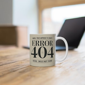 May include: White ceramic mug with the text "SMALL TALK WITHOUT A SCRIPT? ERROR 404 SOCIAL SKILLS NOT FOUND" in black. The mug is on a wooden surface with a laptop in the background.