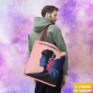May include: A pink tote bag featuring a graphic design of two faces, one blue and one red, with the text "Every day is an emotional plot twist." The bag has a burgundy strap.